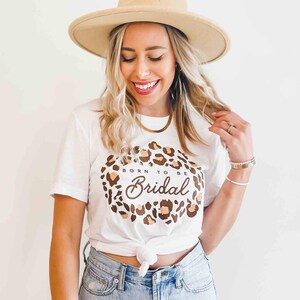 Op de afbeelding: Wit T-shirt met een rond ontwerp van een bruine en beige luipaardprint. De tekst "Born to be Bridal" is in bruin cursief geschreven in de cirkel.