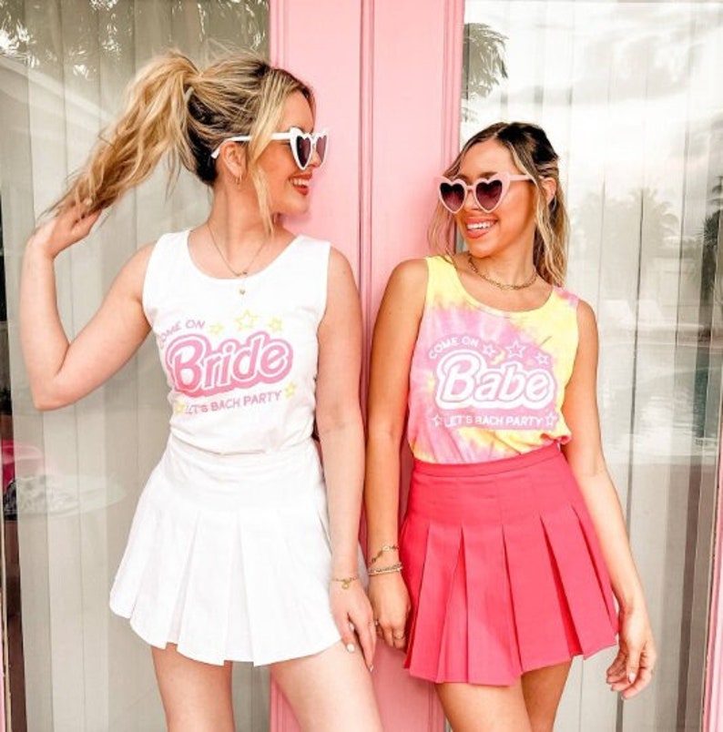 Puede incluir: Dos mujeres con gafas de sol en forma de coraz&oacute;n y conjuntos a juego. Una lleva una camiseta sin mangas y una falda blancas con el texto "Come on Bride". La otra lleva una camiseta sin mangas te&ntilde;ida anudada y una falda rosa con el texto "Come on Babe".