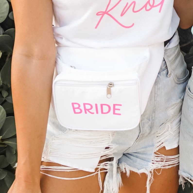 May include: A white fanny pack with the word "BRIDE" in pink lettering. The fanny pack has a zippered front pocket and a silver zipper. The person is wearing light blue denim shorts.