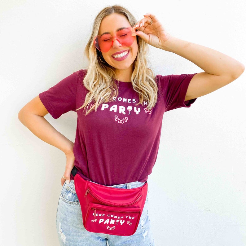 Op de afbeelding: Een bordeauxrood t-shirt met de tekst "HERE COMES THE PARTY" en een rode heuptas met de tekst "HERE COMES THE PARTY". De persoon draagt hartvormige zonnebrillen.