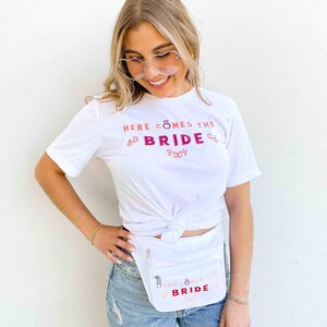 Op de afbeelding: Wit T-shirt en heuptas met de tekst "HERE COMES THE BRIDE" in roze en rood. De heuptas is wit met een ritssluiting. Het shirt is in de taille geknoopt.