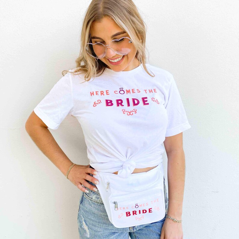 Op de afbeelding: Wit T-shirt en heuptas met de tekst "HERE COMES THE BRIDE" in roze en rood. De heuptas is wit met een ritssluiting. Het shirt is in de taille geknoopt.