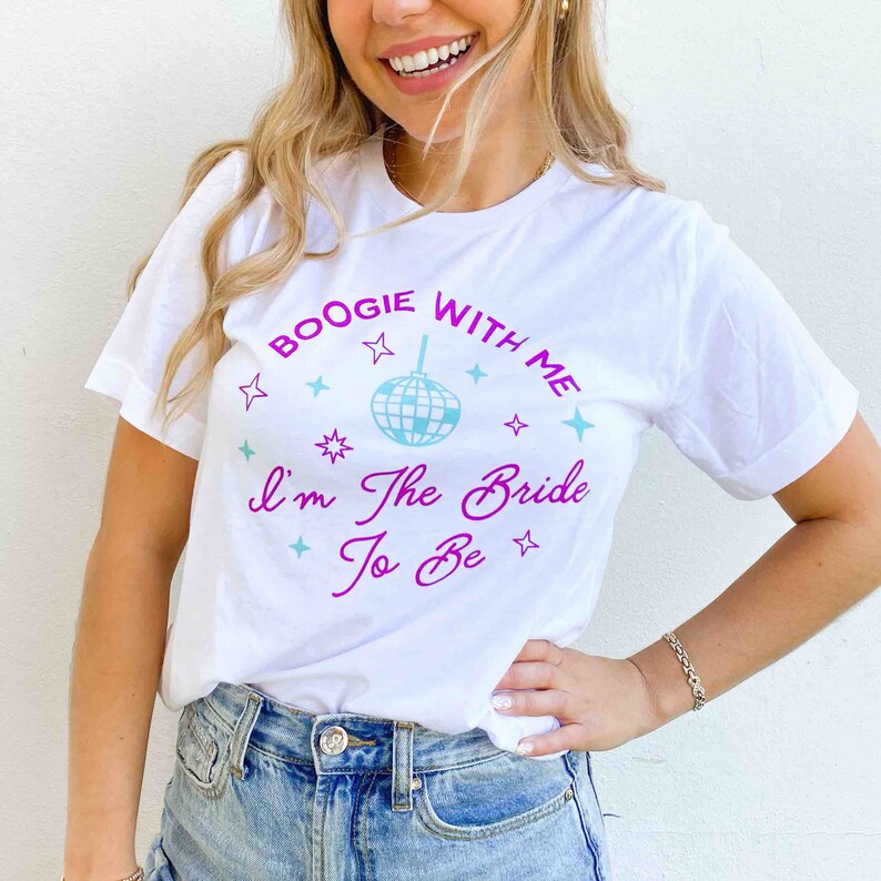 May include: White t-shirt with the text "Boogie With Me I'm The Bride To Be" in a purple and teal design. The design includes a disco ball and star accents. The shirt is a casual, short-sleeved style.