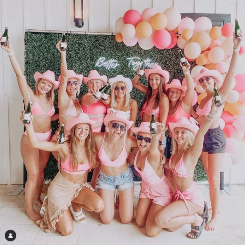 May include: A group of women wearing pink cowboy hats and pink swimsuits pose for a photo in front of a green wall with the words "Better Together" in neon lights. They are holding champagne bottles and smiling.