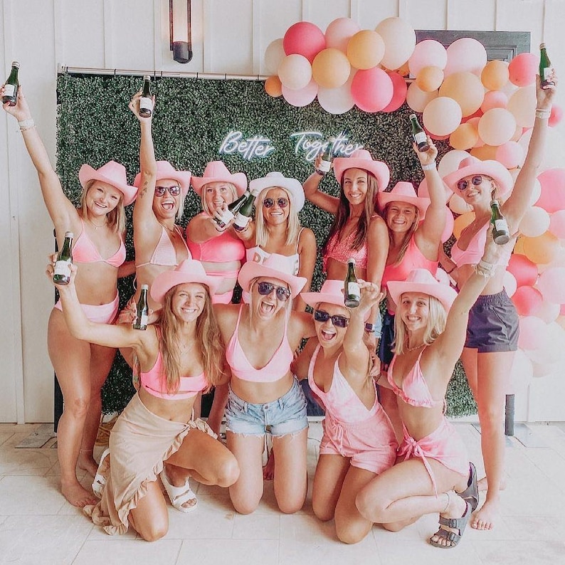 May include: A group of women wearing pink cowboy hats and bikinis pose together in front of a green wall with a neon sign that reads "Better Together". They are holding bottles of champagne and smiling.