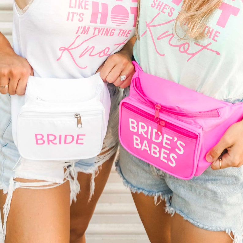 May include: Two fanny packs, one white and one pink, are displayed. The white pack has the word "BRIDE" in pink, while the pink pack says "BRIDE'S BABES" in white. Both have a zippered main compartment and a front pocket. The image suggests a bridal theme.