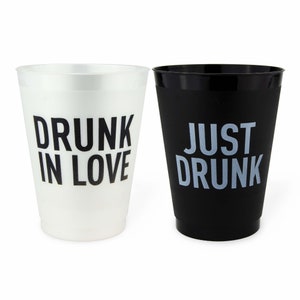 May include: Two plastic cups, one white and one black. The white cup has the text "DRUNK IN LOVE" in black, while the black cup says "JUST DRUNK" in light gray. Ideal for parties and events.