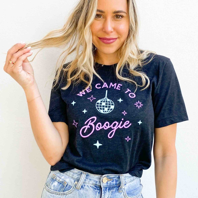 May include: Dark gray t-shirt with the text "WE CAME TO Boogie" in pink, along with a disco ball graphic and star accents. The shirt is made of a soft material and is suitable for casual wear.