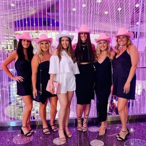 May include: Six women wearing pink cowboy hats and black dresses pose in front of a sparkling wall. One woman is wearing a white dress with a sash that says "Bride to Be".