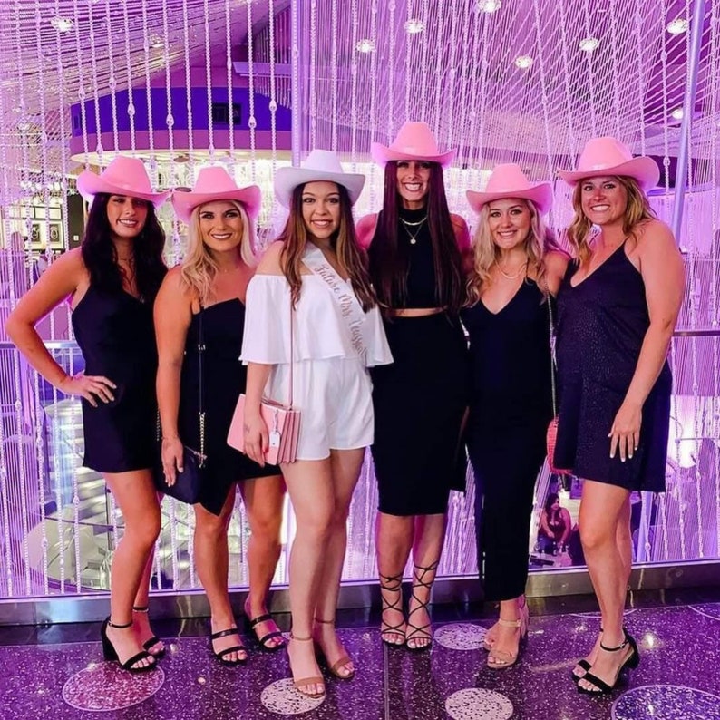 May include: Six women wearing pink cowboy hats and black dresses pose in front of a sparkling wall. One woman is wearing a white dress with a sash that says "Bride to Be".