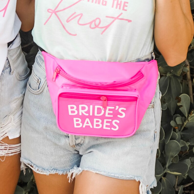 May include: Bright pink fanny pack with the text "BRIDE'S BABES" in white. The fanny pack has a zippered main compartment and a smaller front pocket. The person is wearing denim shorts.
