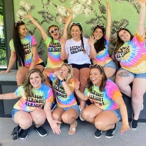 May include: A group of friends wearing tie-dye shirts with the text "Dazed & Engaged" and "Alright Alright Alright". The shirts are in various colours, including pink, blue, yellow, and green. The friends are smiling and posing for a photo.