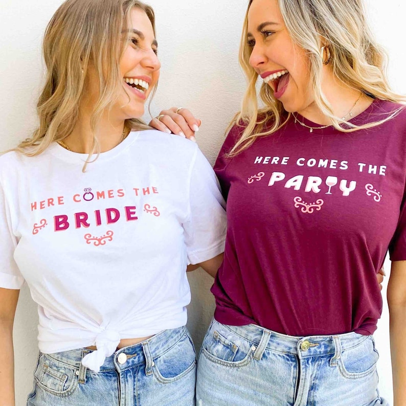 Op de afbeelding: Twee vrouwen dragen bijpassende t-shirts. E&eacute;n is wit met "HERE COMES THE BRIDE" in roze en rood. De andere is bordeauxrood met "HERE COMES THE PARTY" in wit en roze. Beide shirts hebben decoratieve versieringen.