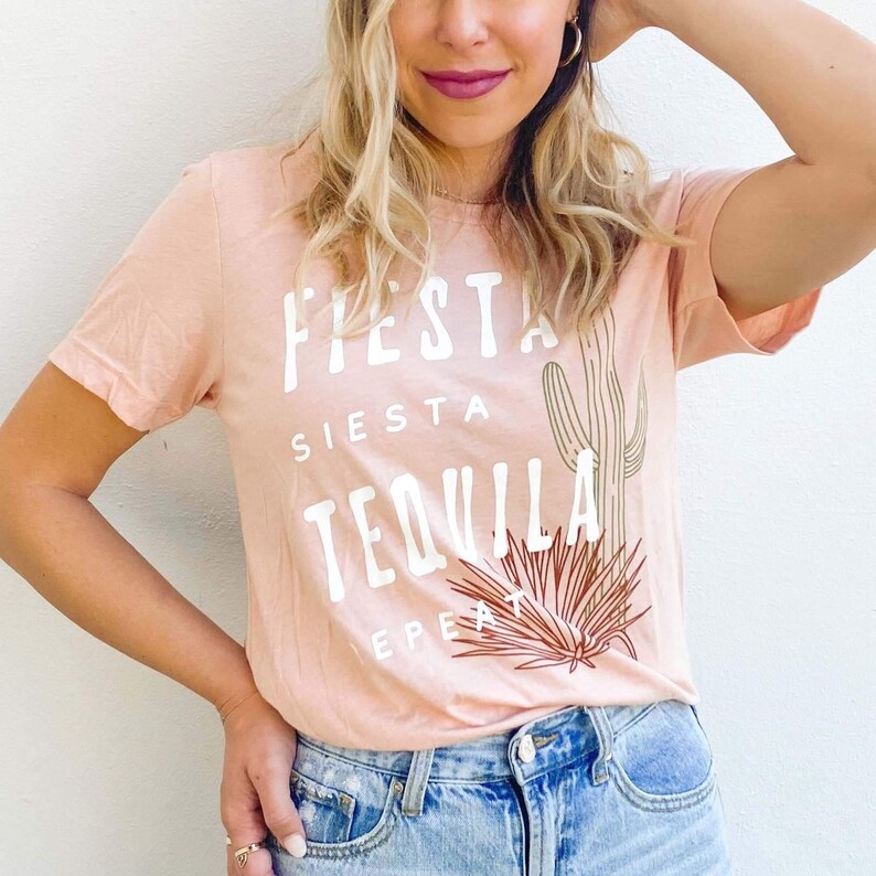 May include: A light peach-colored t-shirt with the words "FIESTA, SIESTA, TEQUILA, REPEAT" in white block letters. A brown cactus and plant graphic are printed below the text. The shirt is paired with blue jeans.