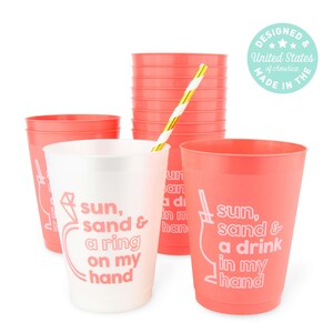 May include: A collection of coral-colored plastic cups with white text that reads "sun, sand & a ring on my hand" and "sun, sand & a drink in my hand." A gold and white striped straw is in one cup. The design is made in the United States.