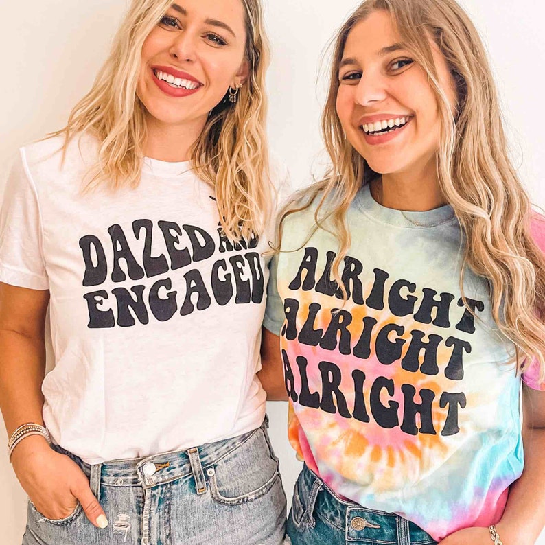 May include: Two women wearing white and tie-dye t-shirts with text. The white shirt says "Dazed and Engaged" in a retro font. The tie-dye shirt says "Alright Alright Alright" in a retro font.