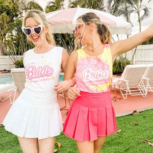 Puede incluir: Dos mujeres con atuendos a juego para una despedida de soltera. Una lleva una camiseta sin mangas y una falda blancas con el texto "Come on Bride, Let's Bach Party". La otra lleva una camiseta sin mangas te&ntilde;ida con tinte y una falda rosa con el texto "Come on Babe, Let's Bach Party".