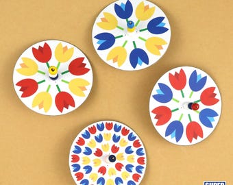 May include: Four colorful paper spinning tops with a tulip design. The tops are red, blue, yellow, and white. The tops are on a brown surface.