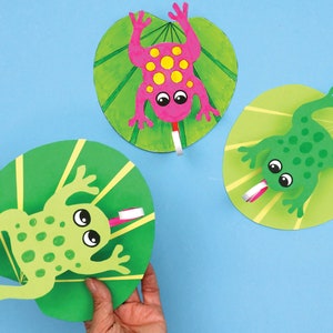 May include: Three paper frog crafts on green lily pads. The frogs have large eyes and are colored green, pink, and green with yellow spots. The frogs have long tongues that are attached to the lily pads with a tab.