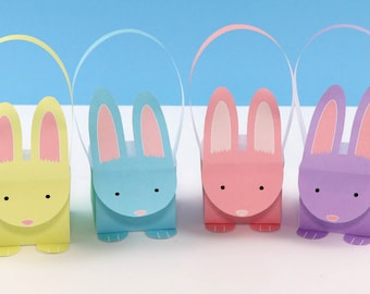 Puede incluir: Cuatro bolsas de regalo de papel con forma de conejo con asas. Las bolsas son amarillas, azules, rosas y moradas. Cada bolsa tiene una cara de conejo con orejas rosas y ojos negros.