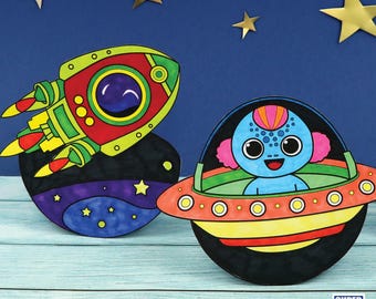 May include: A colorful paper craft set featuring a rocket ship and a UFO with an alien inside. The rocket ship is red, green, and yellow, and the UFO is orange, yellow, and blue. The alien is blue with pink hair and big eyes.