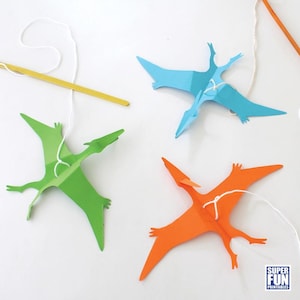 May include: Three paper pterodactyl toys hanging from strings. The pterodactyls are green, blue, and orange. The string is attached to a wooden stick.