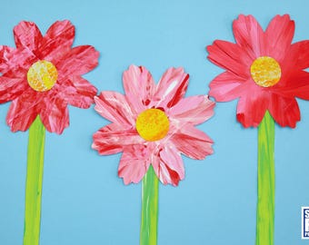 May include: Three paper flowers with pink and red petals and yellow centers. The flowers are on green stems against a blue background.