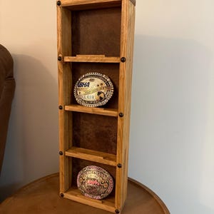 May include: A wooden display shelf with three levels, showcasing two decorative belt buckles. The shelf has a natural wood finish and a brown backing. The belt buckles have intricate designs and text, including "NBHA" and "Summer Series".