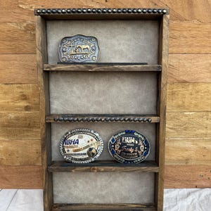 May include: A rustic, wooden display shelf with four levels, adorned with small, silver-colored studs along the top and two shelves. The shelf holds three decorative belt buckles, including one with a horse design and two with text.
