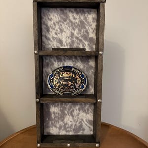 May include: A dark stained wooden display shelf with three levels. The back panel features a cowhide pattern. A decorative belt buckle with the text "STATE LINE 2023 Frosty 5 3D YOUTH CHAMPION" is displayed on the middle shelf.