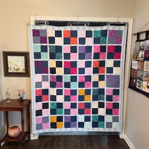 May include: A patchwork quilt featuring a grid of alternating solid dark navy blue squares and various colorful patterned squares in shades of pink, purple, orange, yellow, green, and red, finished with a light blue floral patterned border.
