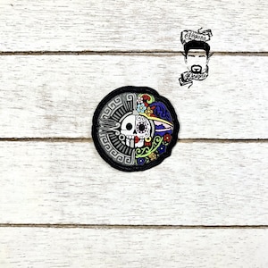 May include: A black and white embroidered patch with a skull and floral design. The skull is surrounded by a colorful floral pattern with yellow, red, blue, and green flowers. The patch has a black border with a decorative pattern.