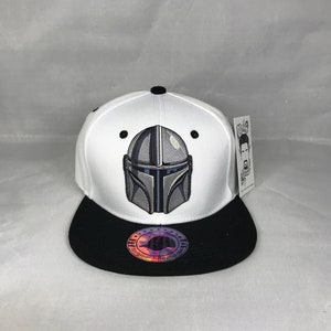 May include: White and black baseball cap with a gray and blue Mandalorian helmet graphic on the front. The cap has a black brim and a white crown. The back of the cap has a black label with the text "Original Snapback".