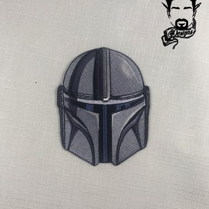 May include: A gray and black embroidered patch featuring a Mandalorian helmet design. The patch has a detailed, textured appearance with dark blue accents. The patch is on a white background.