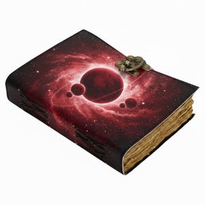 Op de afbeelding: Een leren notitieboek met een ruimtethema. De omslag toont een rode melkweg met planeten en een planeet met ringen. Het heeft een metalen sluiting en verouderde pagina's. Het thema van het notitieboek is ruimte en fantasie.