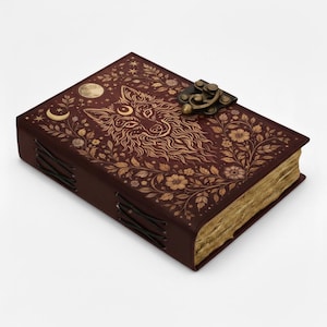 Könnte beinhalten: Ein dunkelbraunes Lederjournal mit Wolfsmotiv und floralen Akzenten. Das Cover zeigt eine detaillierte Illustration eines Wolfsgesichts, eines Mondes und Sterne. Das Journal hat einen Metallverschluss und eine genähte Bindung.