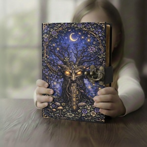 Könnte beinhalten: Ein Fantasy-Tagebuch mit einer detaillierten Illustration eines Baumes mit Gesicht, Mond und Sternen auf dem Einband. Das Tagebuch hat einen Metallverschluss und wird von einer Person gehalten.