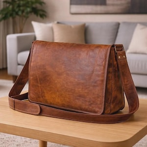 Könnte beinhalten: Braune Leder-Umhängetasche mit Klappe und verstellbarem Schulterriemen. Die Tasche liegt auf einem hellbraunen Holztisch. Die Tasche hat einen Used-Look und befindet sich in einem Wohnzimmer.
