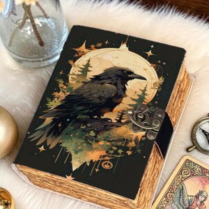 Puede incluir: Un diario decorativo con una ilustración de un cuervo en la portada. El cuervo está posado frente a una luna llena y una escena de bosque. El diario tiene un cierre de metal y páginas envejecidas. Se ve una carta del tarot y una pequeña brújula.