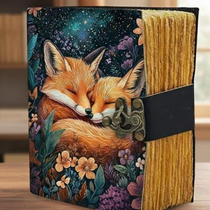 Könnte beinhalten: Ein dekoratives Notizbuch mit einem Einband, der zwei orange Füchse zeigt, die sich inmitten von Blumen und einem Sternenhimmel umarmen. Das Notizbuch hat einen schwarzen Rücken, einen Verschluss und gealterte Papierseiten.
