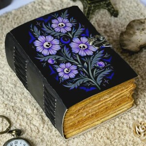 Könnte beinhalten: Ein schwarzes Notizbuch mit einem floralen Design mit lila Blumen mit Totenkopfzentren und blauen Akzenten. Das Notizbuch hat einen Metallverschluss und gealterte Seiten. Der Einband ist aus Leder.