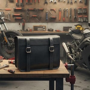 Könnte beinhalten: Eine schwarze Motorrad-Satteltasche aus Leder mit silbernen Schnallen steht auf einer hölzernen Werkbank. Die Tasche hat zwei Riemen und ist rechteckig. Im Hintergrund ist ein Motorrad zu sehen, zusammen mit Werkzeugen, die an einer Lochwand hängen.