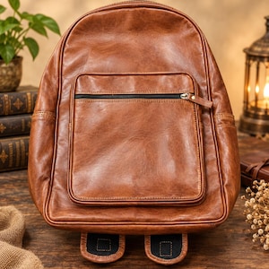 Può includere: Uno zaino in pelle marrone con un ampio scomparto principale e una tasca frontale più piccola con chiusura a zip nera. La borsa presenta una finitura liscia e lucida e due spallacci imbottiti. Il testo LEATHER MART è stampato.