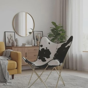 May include: Butterfly chair with a black and white cowhide pattern seat and gold-colored metal frame. The chair is in a living room setting with a yellow armchair, a round mirror, and a wooden cabinet.