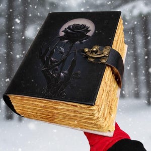 May include: Black leather-bound journal with a black rose design on the cover. The cover features a black rose and a hand reaching for the rose, with a full moon in the background. The pages have a vintage, aged look, and the journal is secured with a leather strap and clasp.