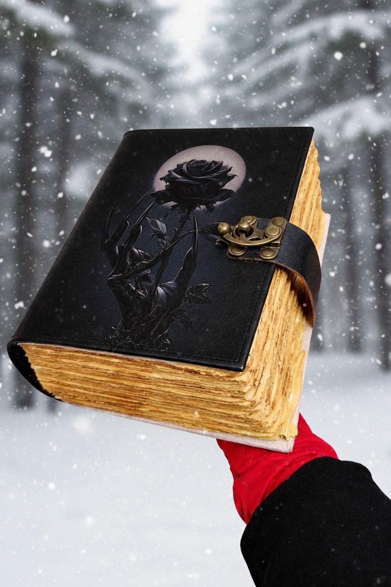 May include: Black leather-bound journal with a black rose design on the cover. The cover features a black rose and a hand reaching for the rose, with a full moon in the background. The pages have a vintage, aged look, and the journal is secured with a leather strap and clasp.