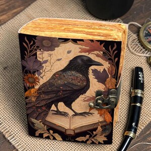 Könnte beinhalten: Ein dekoratives Notizbuch mit einer Rabenillustration auf dem Einband. Der Rabe sitzt auf einem offenen Buch, umgeben von Herbstblättern und Blumen. Das Notizbuch hat einen Verschluss und gealterte Seiten.