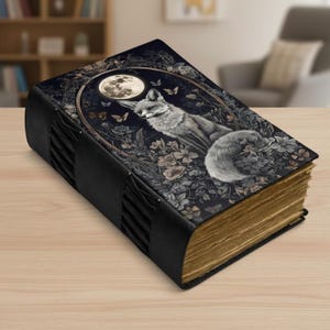 Könnte beinhalten: Ein dekoratives Notizbuch mit schwarzem Ledereinband und einer detaillierten Illustration eines Fuchses unter einem Vollmond. Das Cover-Art zeigt florale und Schmetterlingsakzente. Die gealterten Seiten sind sichtbar, und das Notizbuch ist mit schwarzen Lederriemen gebunden.