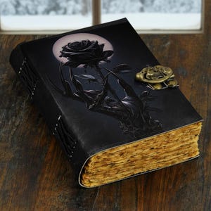 Könnte beinhalten: Schwarzes Lederjournal mit Rosen- und Handdesign. Das Cover zeigt eine schwarze Rose und eine Handgrafik mit einem Vollmond im Hintergrund. Das Journal hat einen Messingverschluss und alt aussehende Seiten. Das Journal ist mit schwarzem Faden gebunden.
