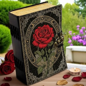 Könnte beinhalten: Ein schwarzes Notizbuch mit einer großen roten Rosenillustration auf dem Einband. Der Einband hat ein verziertes Design mit goldenen Akzenten und einem Verschluss. Die Seiten haben einen Goldrand. Rote Rosenblätter und Kerzen sind in der Nähe.